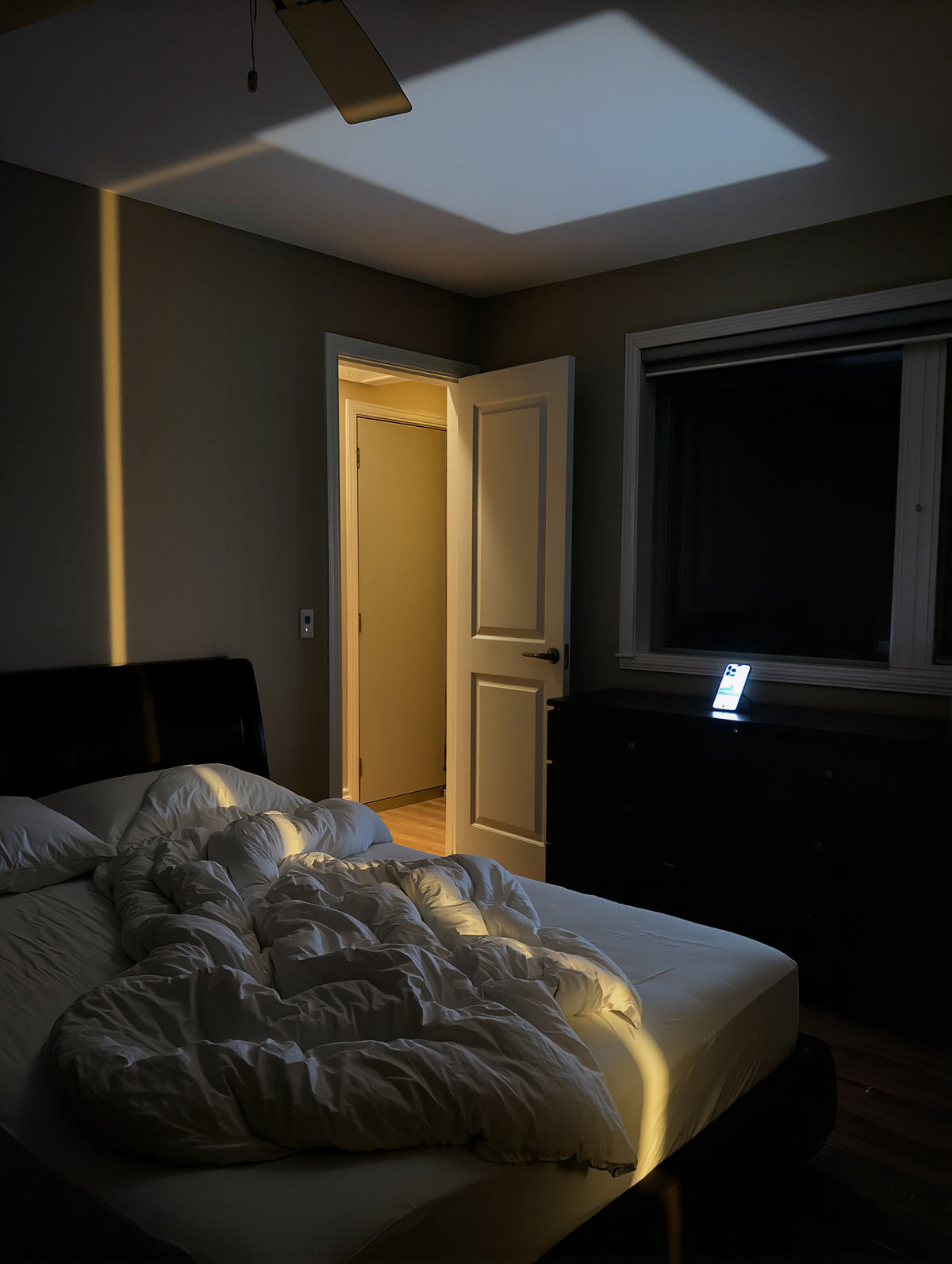 A pitch-dark bedroom at 6:45 with a phone alarm glowing on the dresser across the room and hallway light under the door
